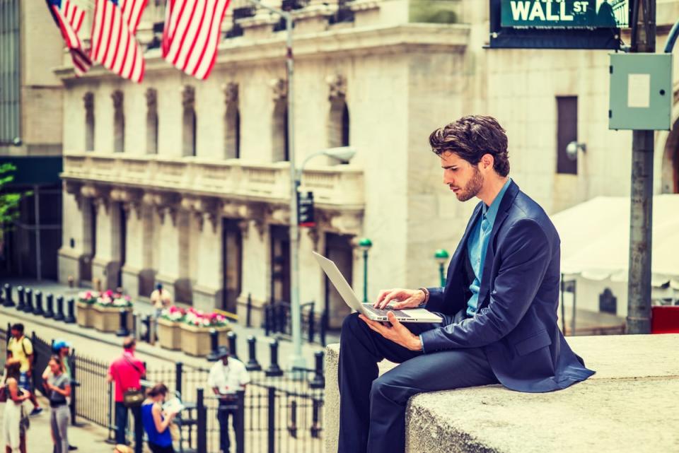 A man sitting on a ledge looks at his laptop.  The street sign above him says Wall St.