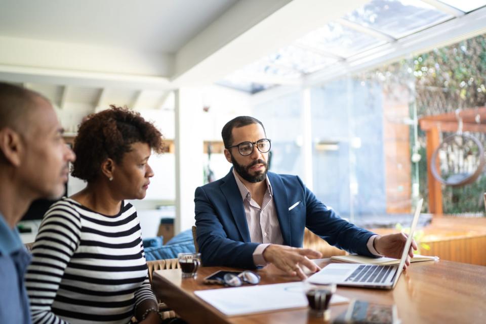 Two people sitting at a table listening to someone explain something to them on a laptop. 