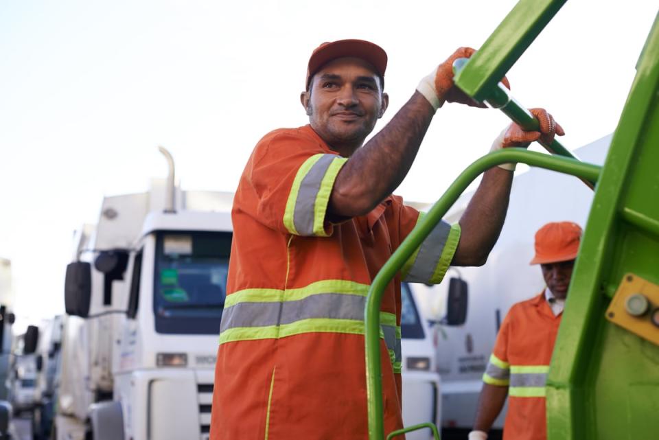 A person holding the back of a garbage truck. 