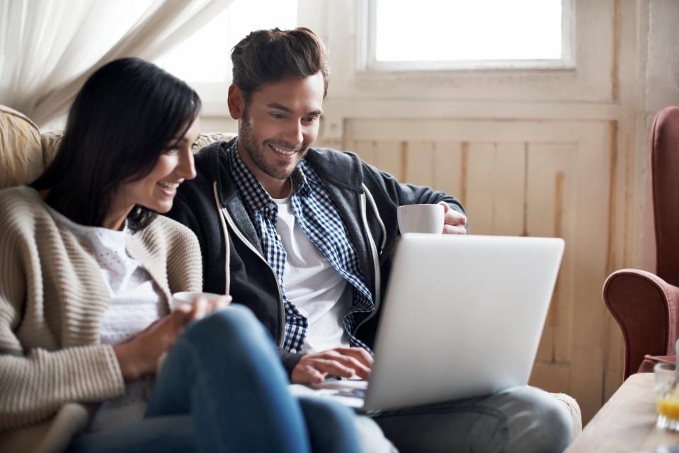 Two people sitting on a sofa smile while looking at something on a laptop.