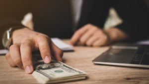 hands at desk near laptop, with one hand holding a stack of one hundred dollar bills.  Banking shares