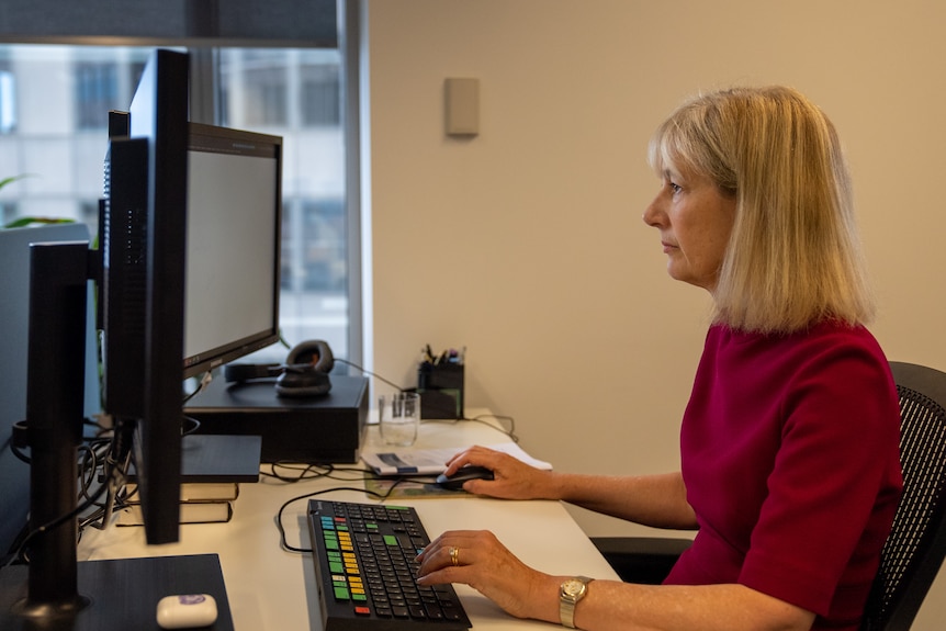 A blonde woman wearing a pink blouse is sitting in front of the computer.
