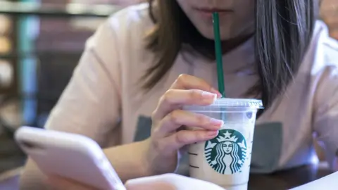 Getty Images Woman sitting at a table drinking cold Starbucks drink through a straw while looking at her cell phone