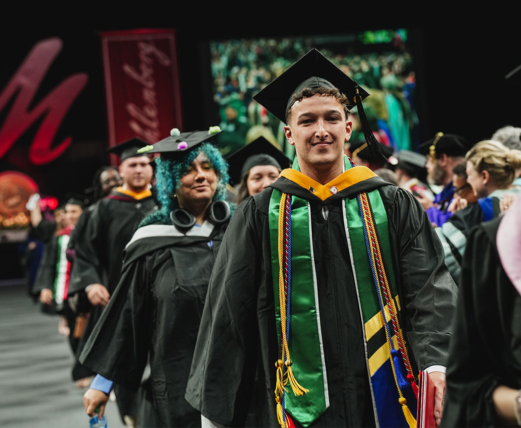 New graduates, smiling in commencement regalia, file out of an arena.