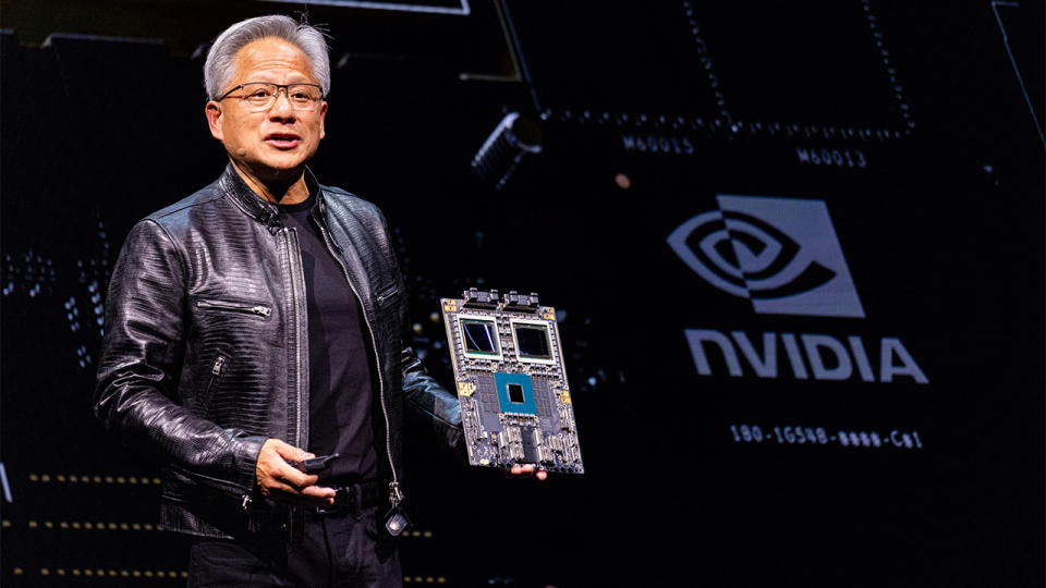 Huang holds up a circuit board while giving a lecture.