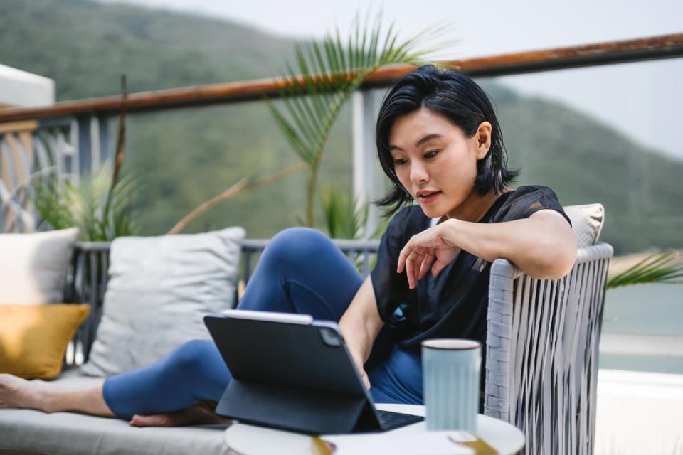 A person lounging on a sofa on a balcony in a coastal setting while interacting with a tablet on a table. 