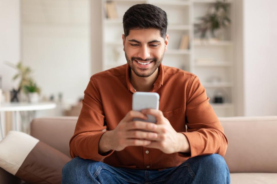 A person in jeans and a button-down shirt sitting on a couch smiling at a phone in their hands.