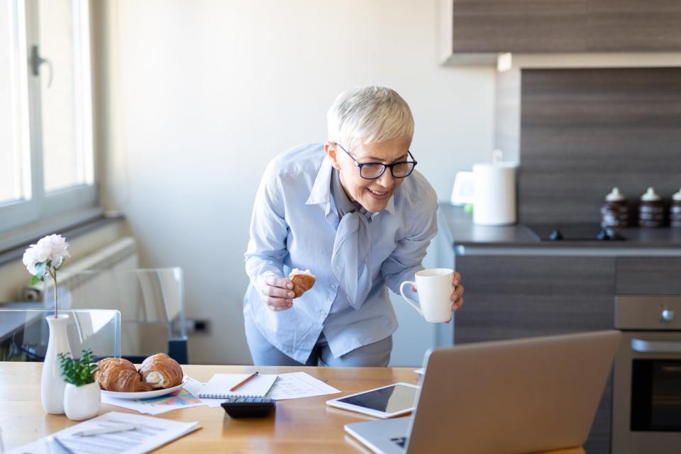 A woman stands in her kitchen holding a cup of coffee while looking at her laptop on a table with a smile on her face